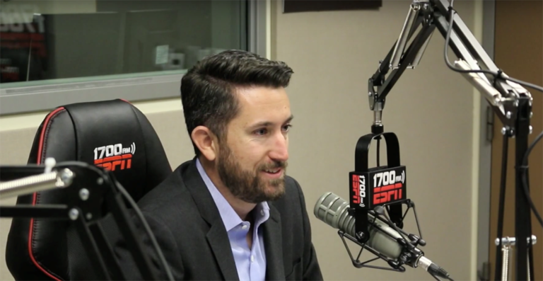 Keynote speaker Dr. Eric Bean is speaking into a microphone at a radio studio. The microphone and chair he is seated on read "1700 ESPN."